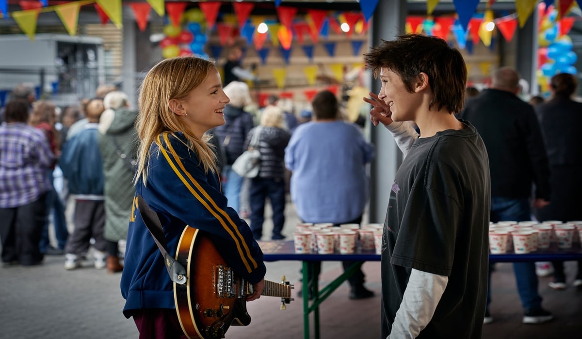 En tjej med elgitarr och en kille står och pratar på en plats med partyflaggor och mycket folk