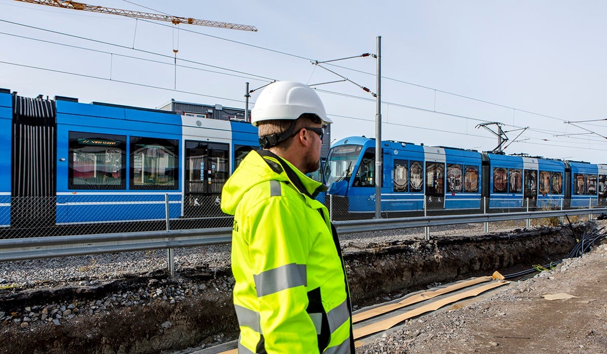 En man iförd skyddshjälm och gul reflexväst står framför ett tåg.