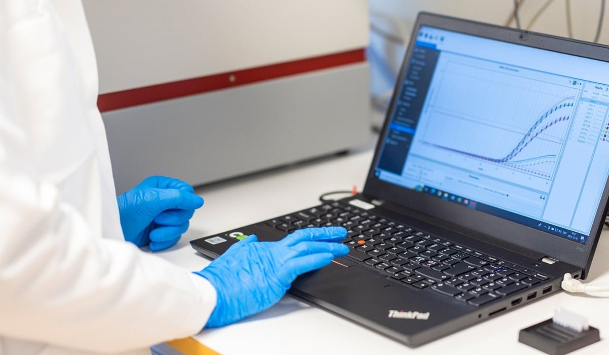 Person i labbkläder och handskar , personen läser av en kurva
 på en dataskärm