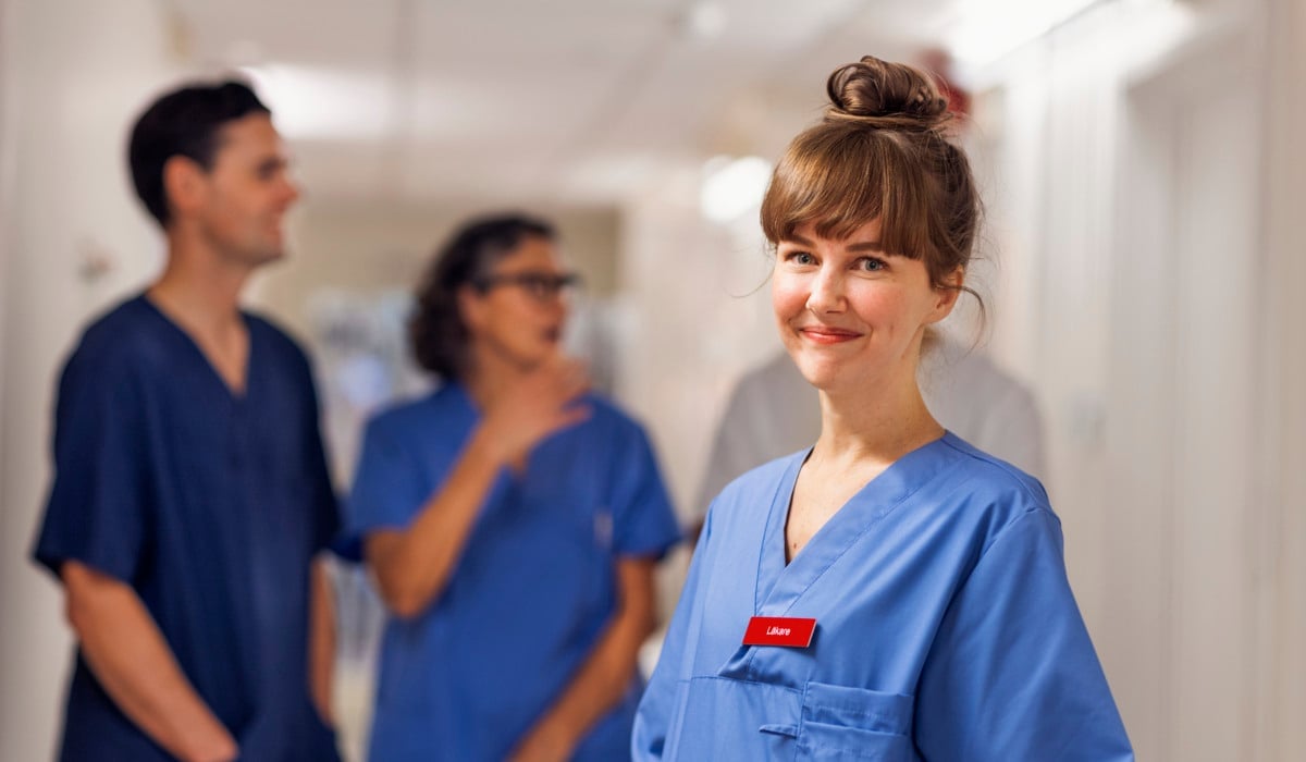 En ung kvinnlig läkare som står i en korridor i sjukhusmiljö. Hon tittar in i kameran och ler. I bakgrunden står tre kollegor till läkaren och pratar med varandra.
