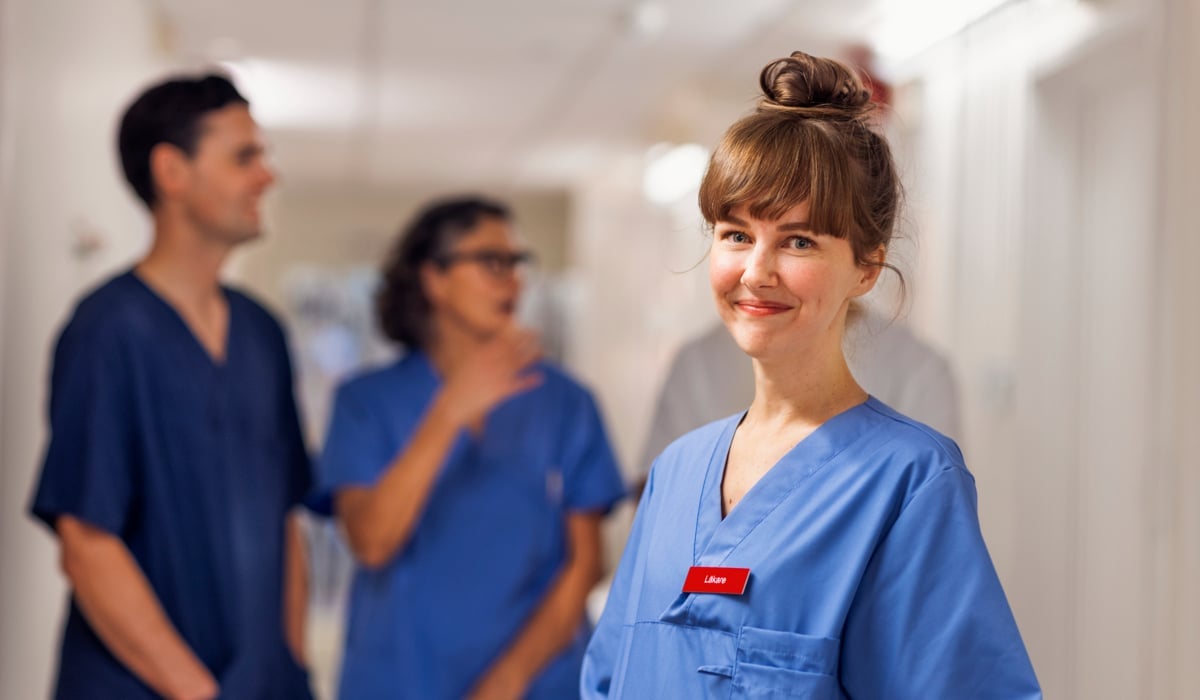 En ung kvinnlig läkare som står i en korridor i sjukhusmiljö. Hon tittar in i kameran och ler. I bakgrunden står tre kollegor till läkaren och pratar med varandra.