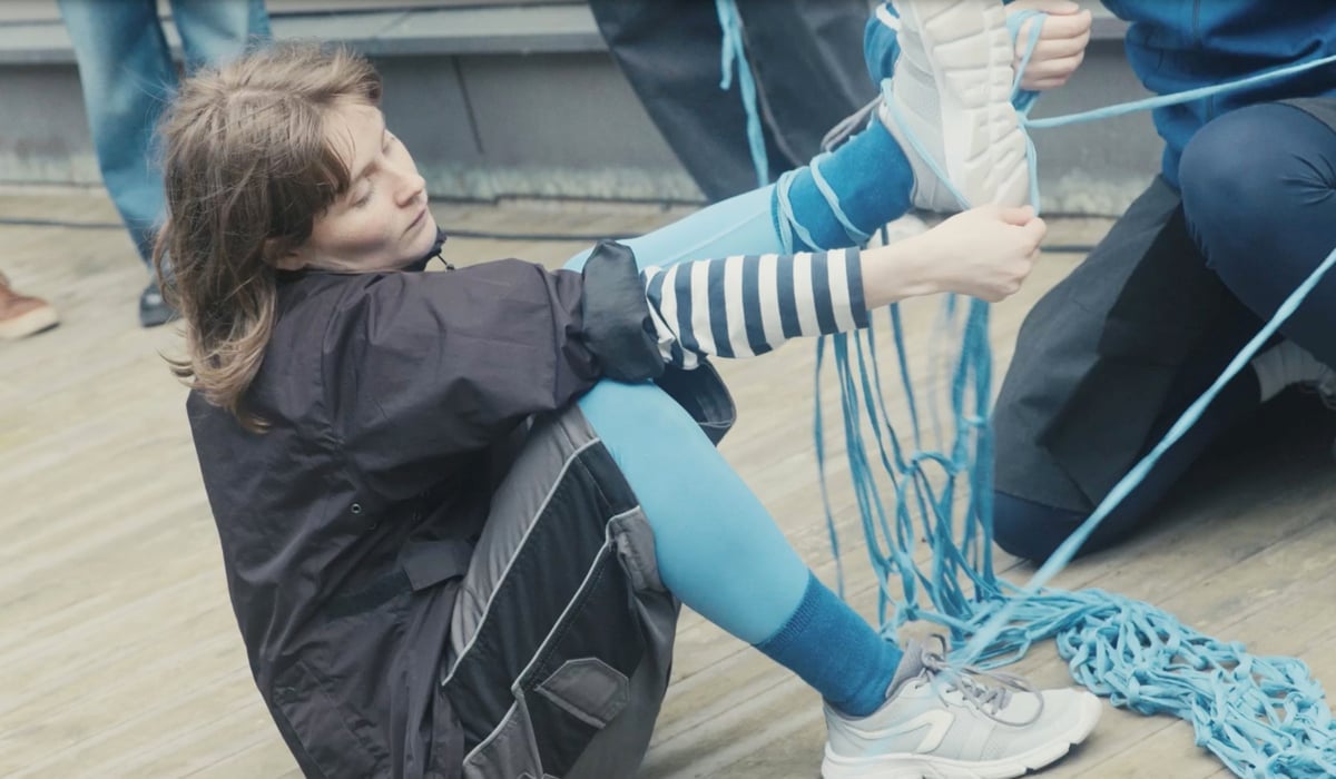 En dansare sitter på marken med benen och fötterna intrasslade i en stor virkning. Hon bär ytterkläder och det blåser lätt i hennes hår. Runt henne skymtar andra ben och fötter.