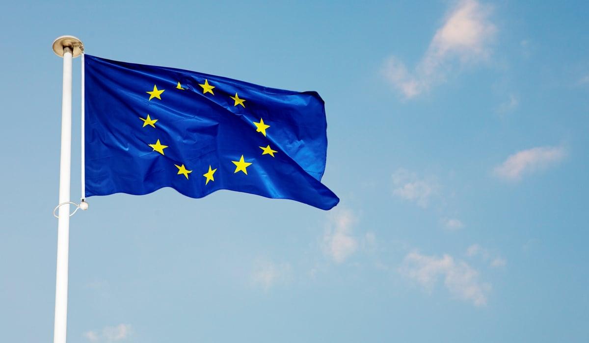 Bild på en blå EU-flagga som vajrar på en vit flaggstång med blå himmel i bakgrunden