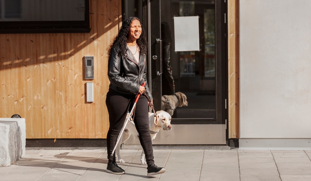 En synskadad kvinna som är ute och tar en promenad. I sin ena hand håller hon sin ledhund, i den andra handen håller kvinnan en blindkäpp