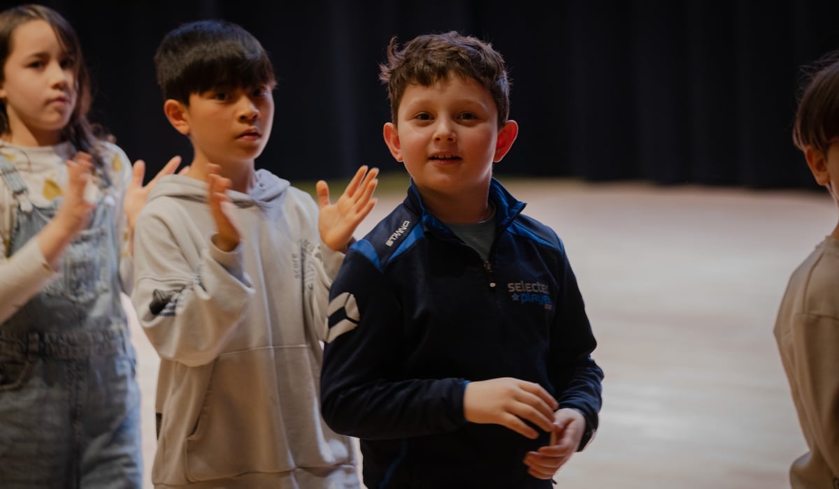 Barn i en danssal. En pojke tittar in i kameran, två klappar händer.