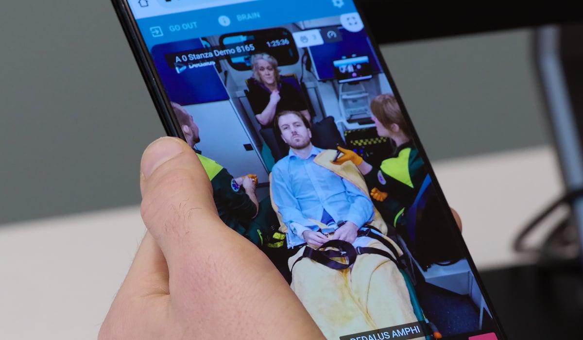 Foto på en hand som håller i en mobiltelefon. På telefonens skärm syns en man som blir omhändertagen av ambulanspersonal. 