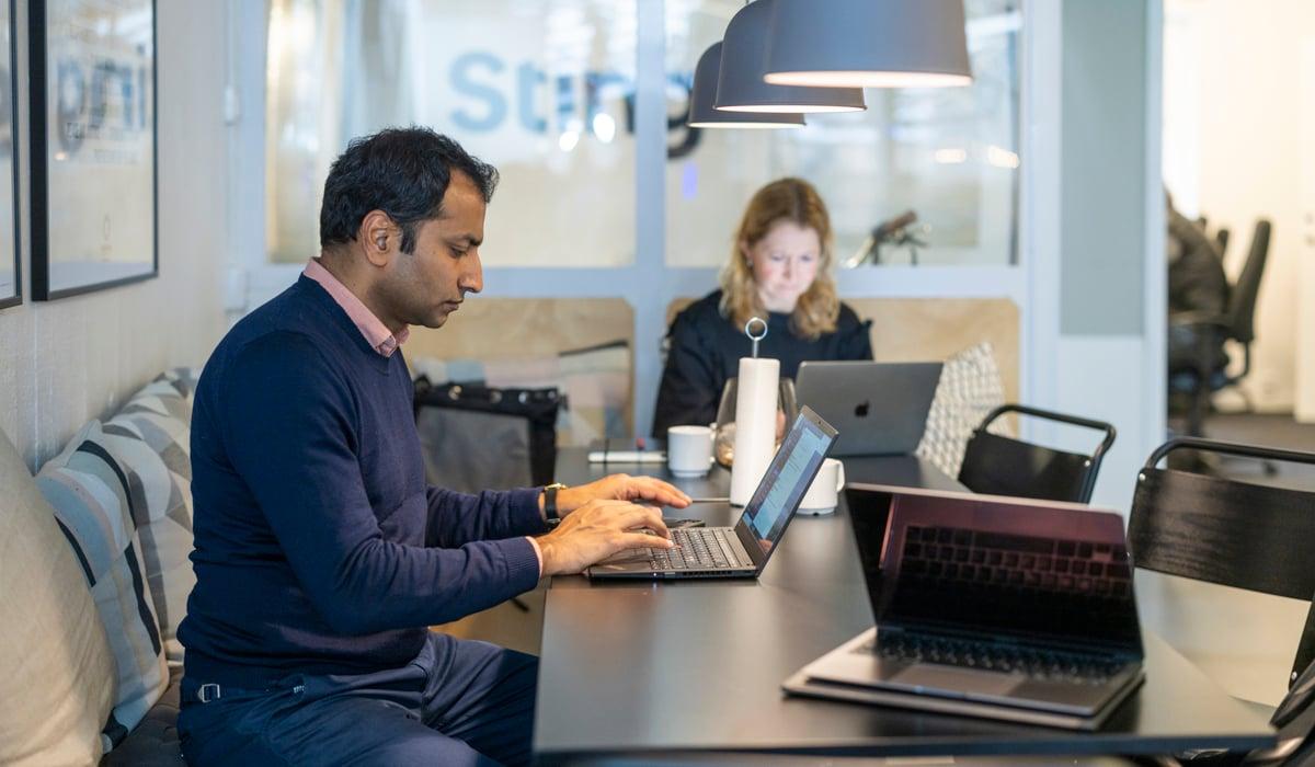 Foto på två personer som sitter vid ett bord med en varsin bärbar dator framför sig. 
