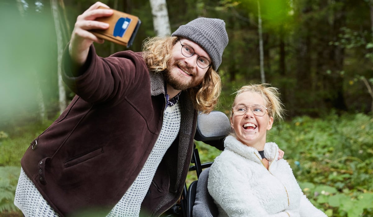 Ung manlig vårdare tar selfie med kvinna i rullstol i skog.