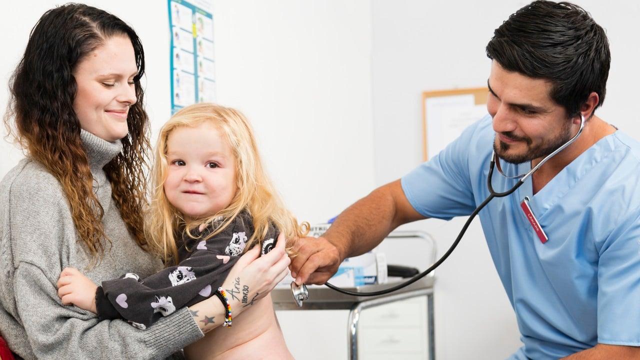 Manlig läkare lyssnar med stetoskop på ryggen på en liten flicka som sitter i sin mammas knä