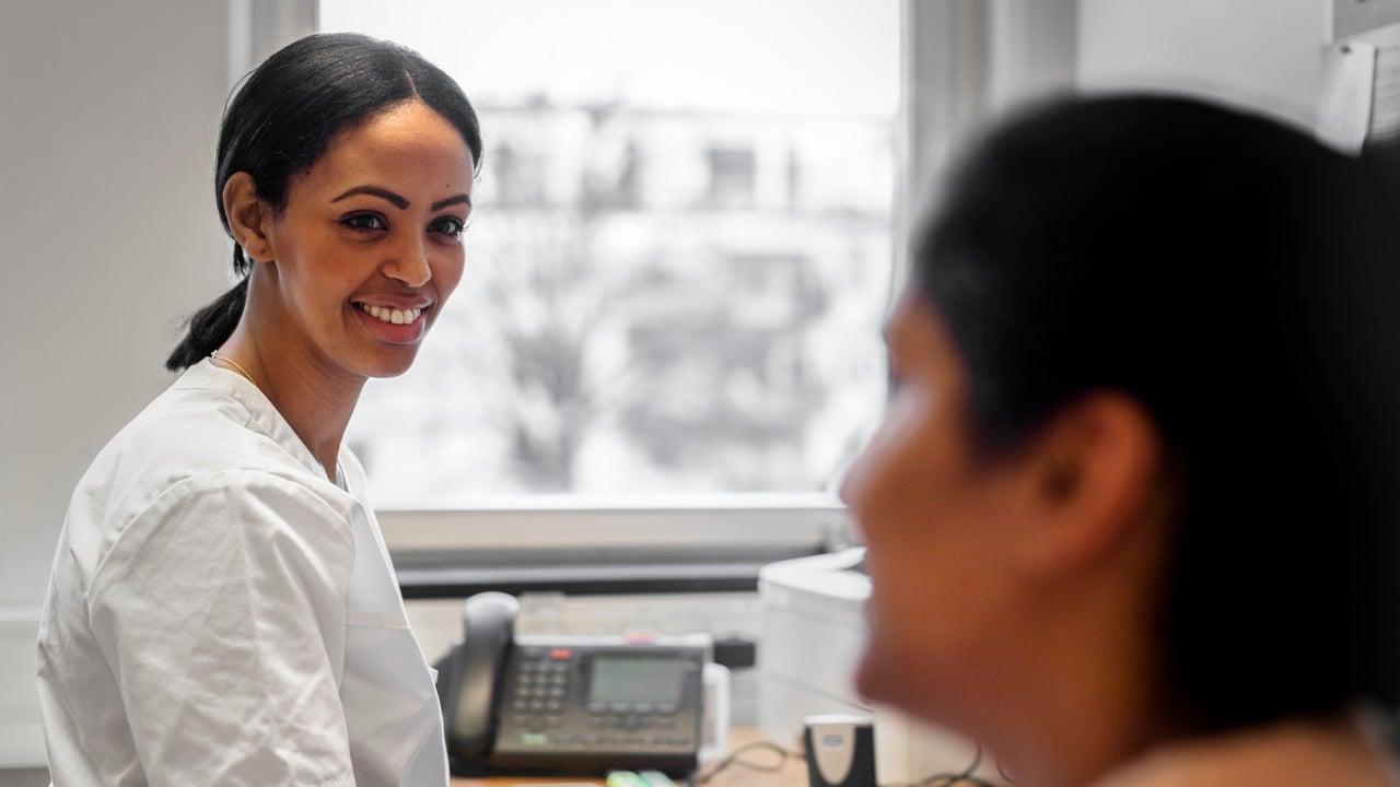 Kvinnlig vårdmedarbetare sitter vid ett skrivbord och tittar på en patient som syns i förgrunden