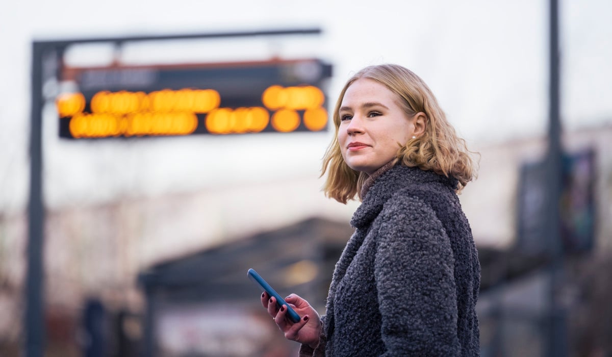 En kvinna står framför en busshållplats med en mobiltelefon i handen.