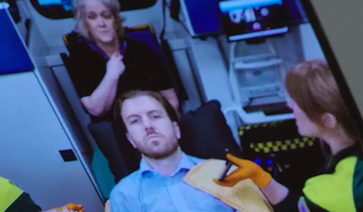 En patient sitter på en bår i en ambulans. Bilden visas på en mobilskärm.