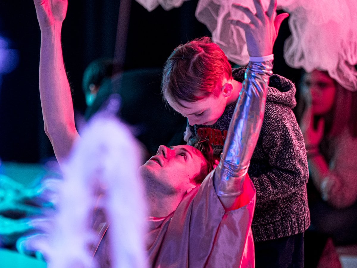 Ett barn står bakom en silverklädd dansare som sitter med armarna uppsträckta i luften. Runt om dem syns olika tylltyger.