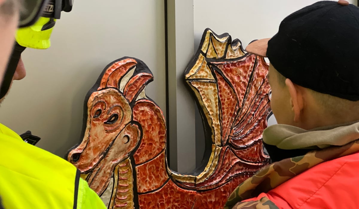 En man i gul jacka och en kvinna i svart mössa framför drake i keramik utomhus.