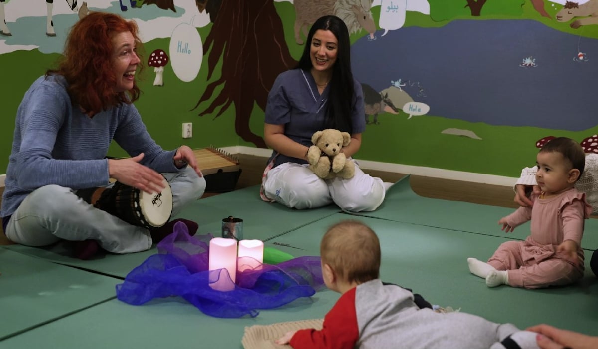 Bebisar på golv tittar på kvinna som ler och spelar handtrumma. I mitten av rummet låtsasljus på tyg. En mysig stämning för bebisarna.