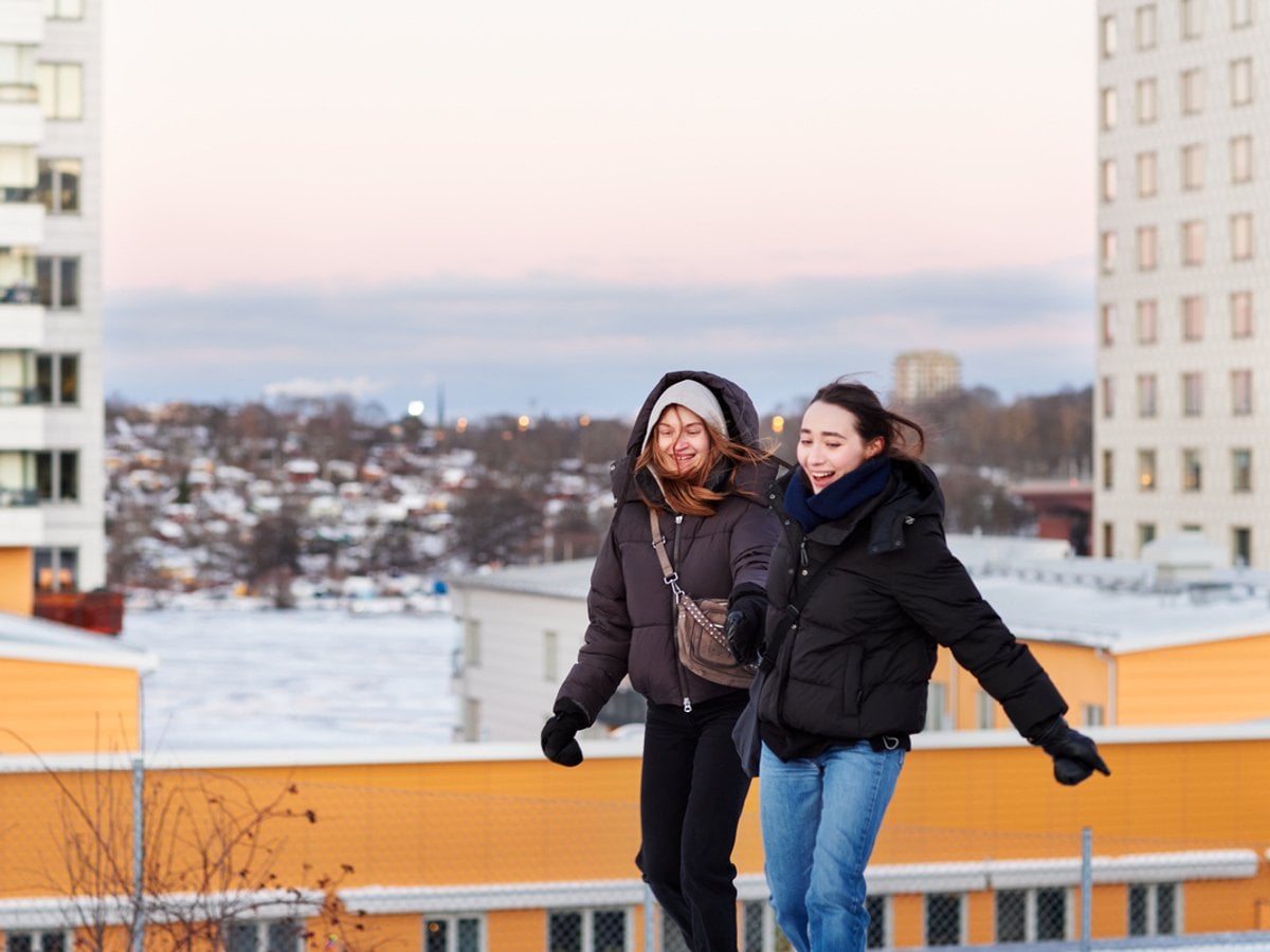 Foto på två kvinnor som går utomhus och håller i varandra. I bakgrunden syns höghus.
