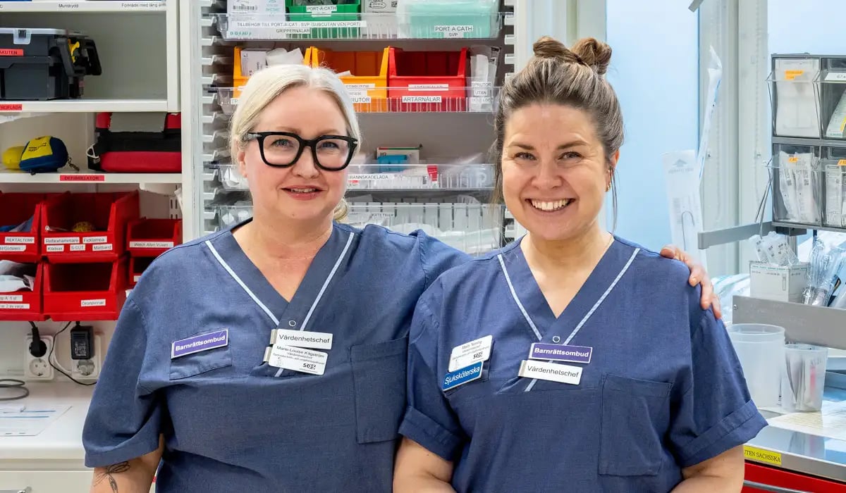 Foto på Marie-Louise och Malin som jobbar som barnrättsombud. De är klädda i sjukhuskläder och ler in i kameran.