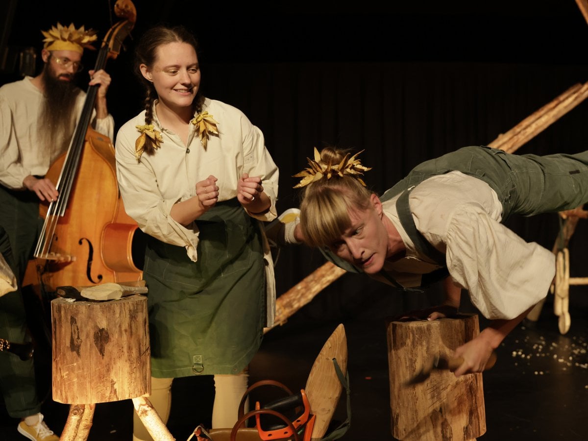 En man med krans på huvudet spelar kontrabas, en kvinna med flätor hejar på och en kvinna i snickarbyxor står på en hand på en huggkubbe-stubbe