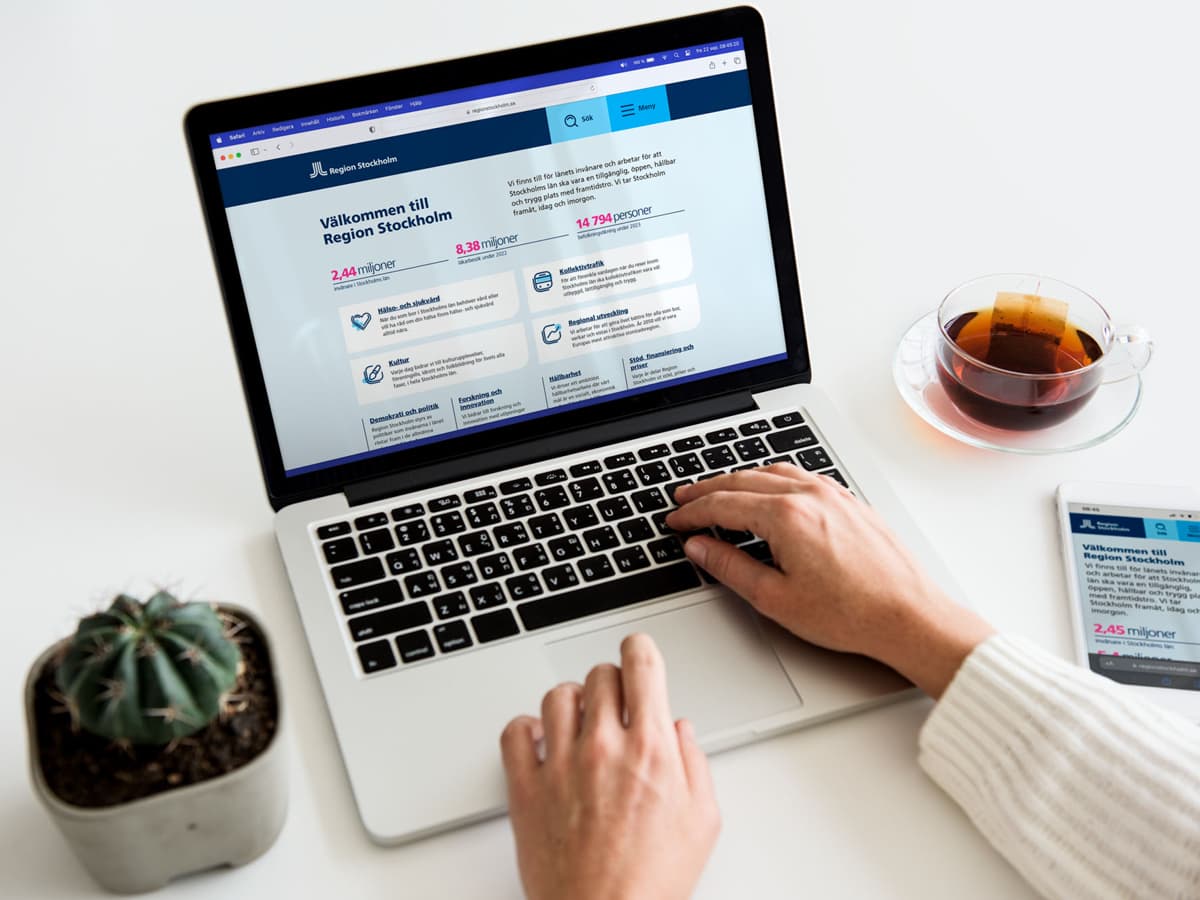 Foto på en person som använder en dator och har en mobiltelefon bredvid datorn. På datorn och mobiltelefon är Region Stockholms webbsida öppen