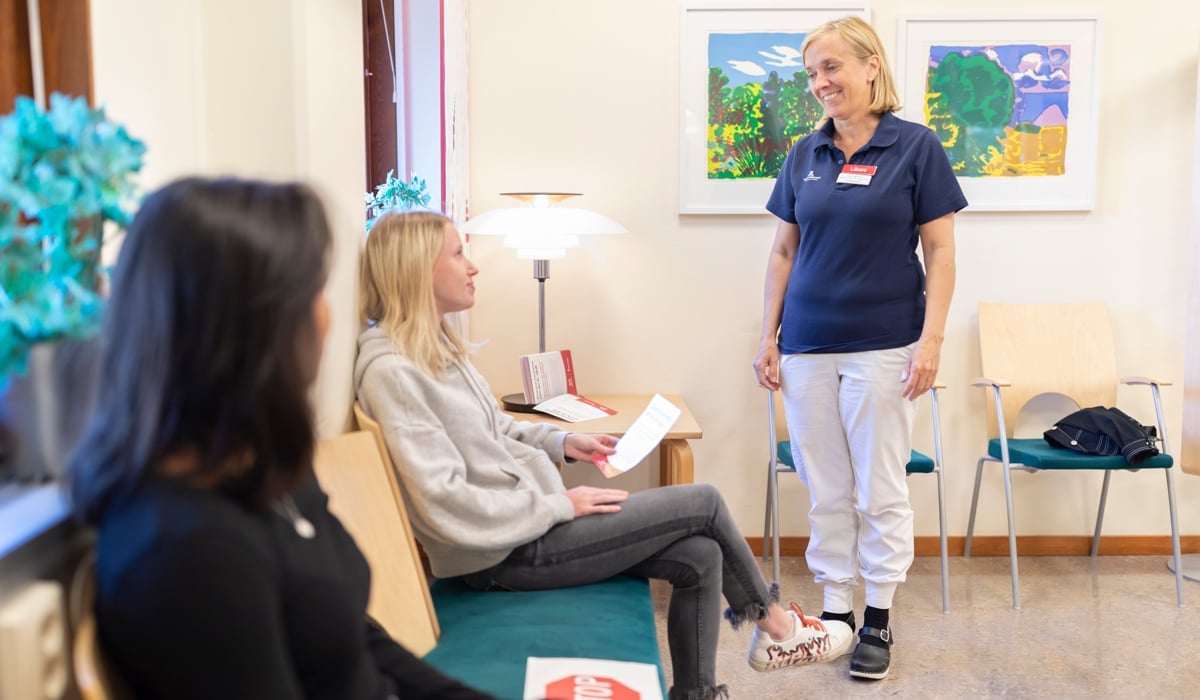 En kvinnlig läkare står i ett väntrum med två väntande patienter.