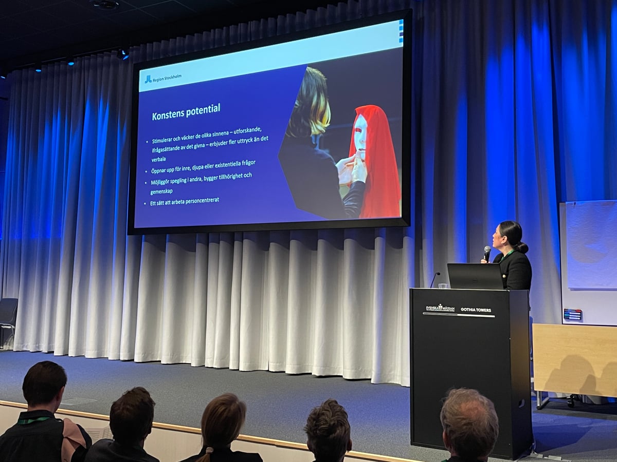 Broula på en scen med åhörare framför sig som alla tittar på stor skärm med presentation.