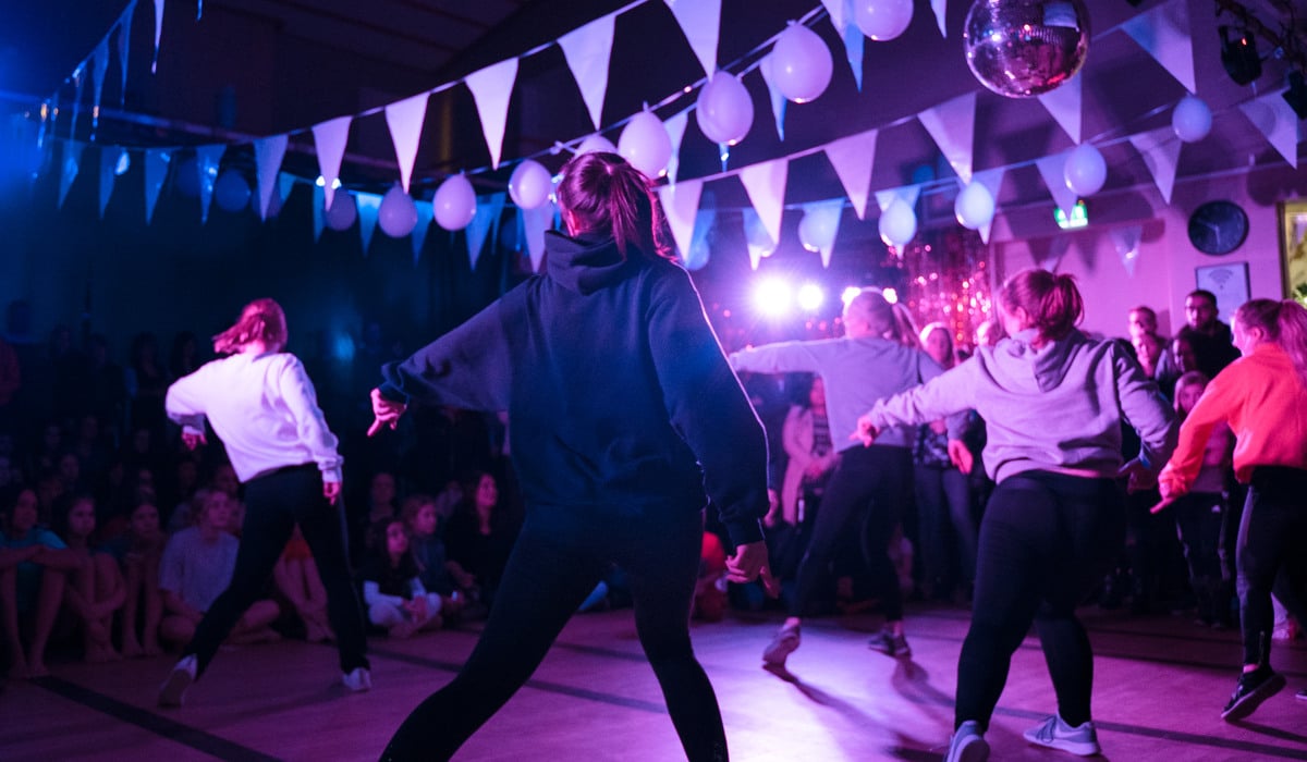 Fem dansare dansar en synkroniserad koreografi på ett dansgolv i en mörk lokal med flaggor, ballonger och festlig belysning i taket.