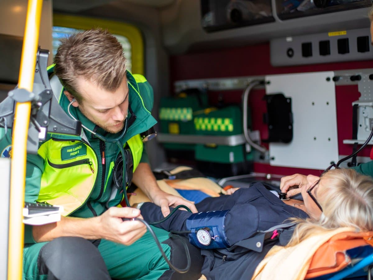 Två medarbetare inom ambulanssjukvården i neongröna arbetskläder tar hand om en kvinnlig patient som sitter på en bår.
