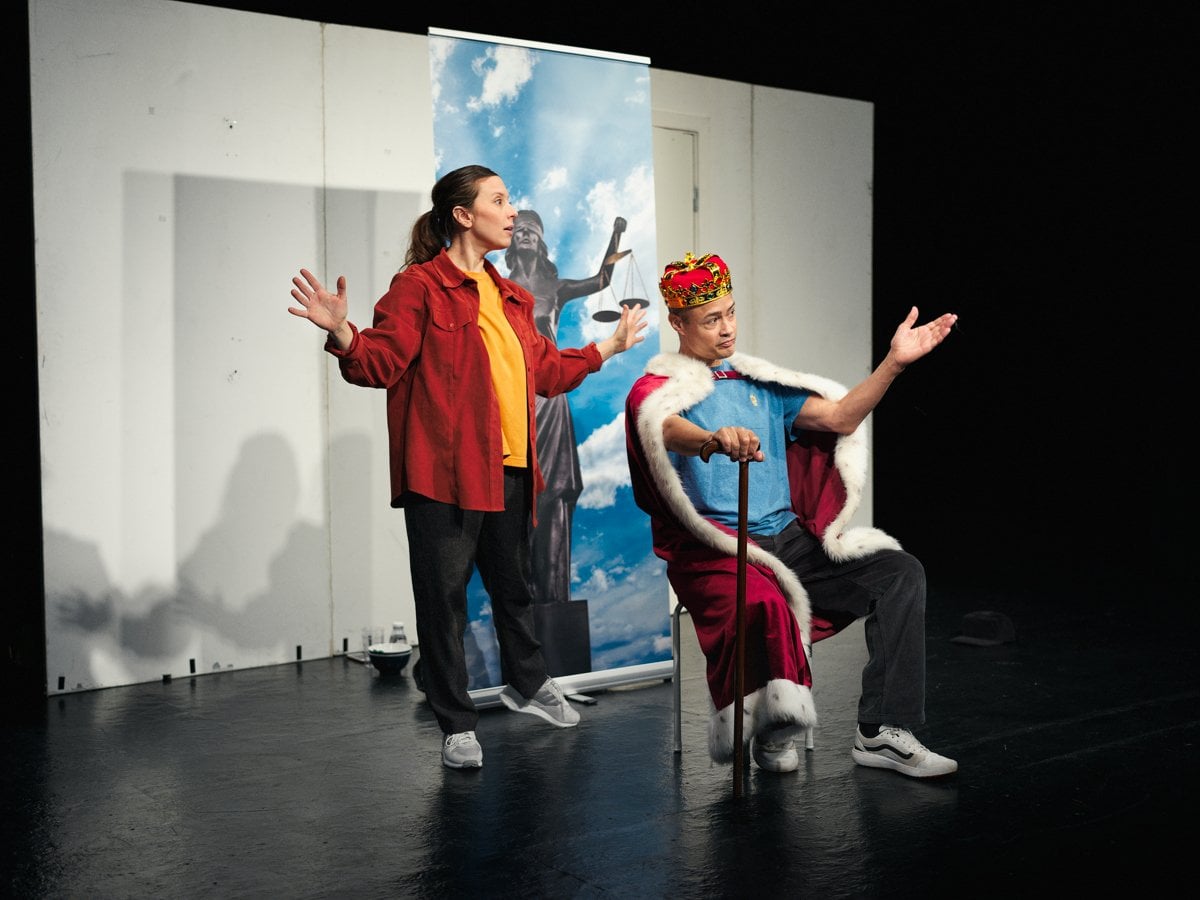 Två skådespelare på scen. En kvinna står med armarna i luften bakom en man som sitter ner med en kungakrona på huvudet och håller i en käpp.