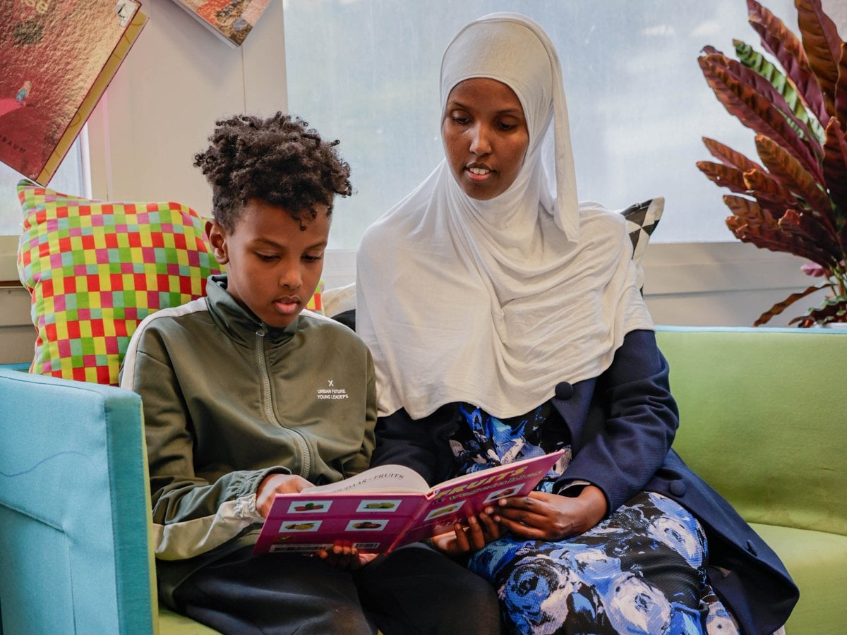En pojke sitter och läser på biblioteket tillsammans med sin mamma i en grön soffa.