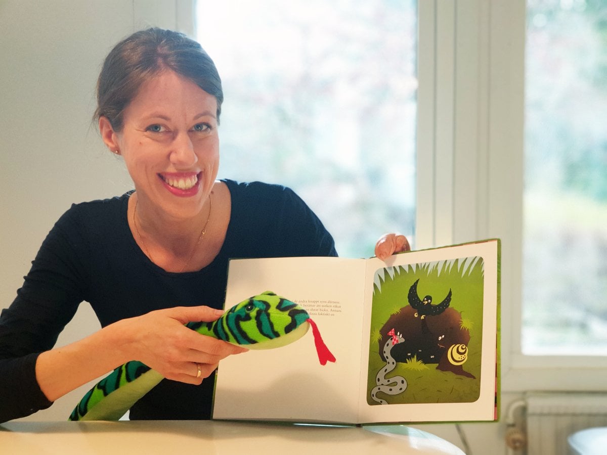 En kvinna sitter med en uppslagen barnbok och håller i en mjukisorm och ler mot kameran.