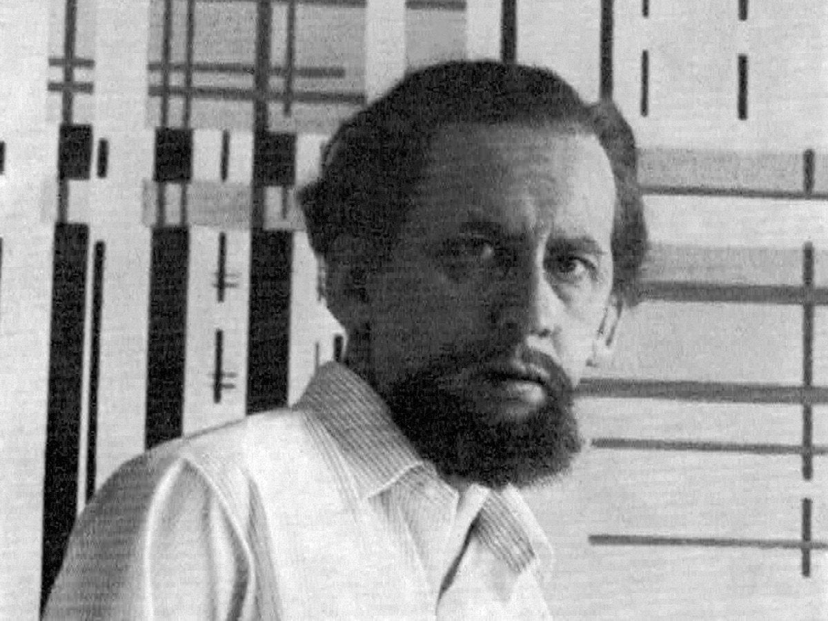 Svart-vitt foto av Reis som tittar mot kamerian iförd randig skjorta mot rutig vägg. Leo har mustigt skägg och långt hår.