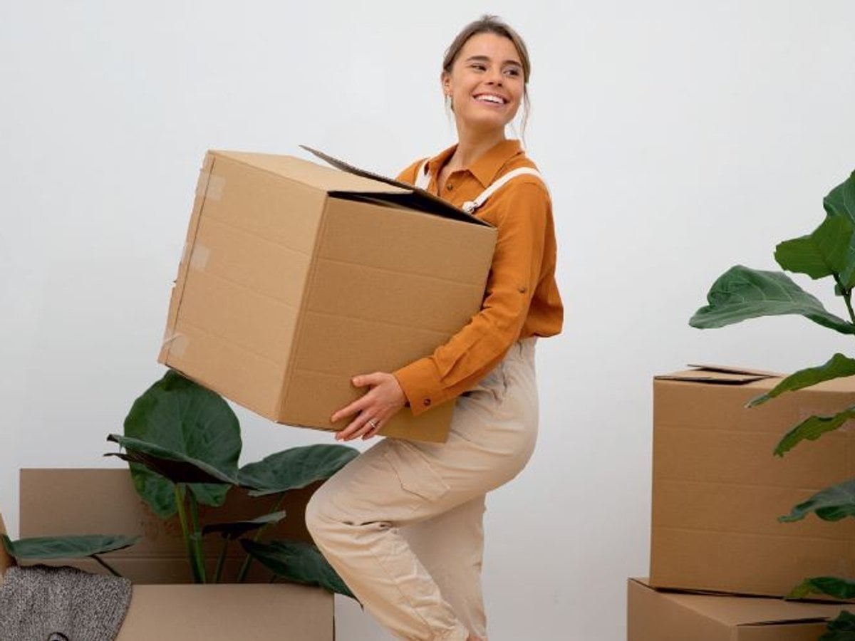 Foto på en kvinna som bär på en flyttlåda. I bakgrunden syns fler flytttådor samt växter.