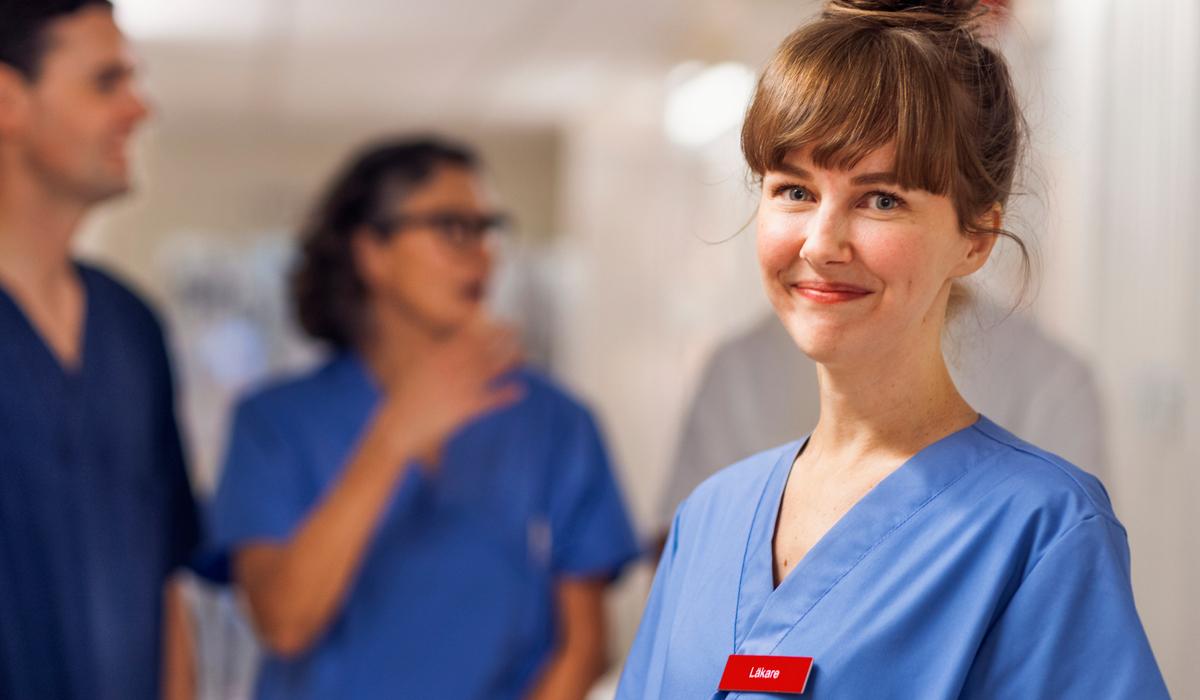 En ung kvinnlig läkare som står i en korridor i sjukhusmiljö. Hon tittar in i kameran och ler. I bakgrunden står tre kollegor till läkaren och pratar med varandra.