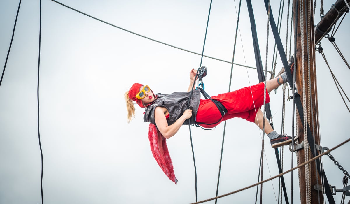 En kvinna som är cirkusartist hänger i ett rep och en sele med fötterna mot masten på ett segelfartyg.