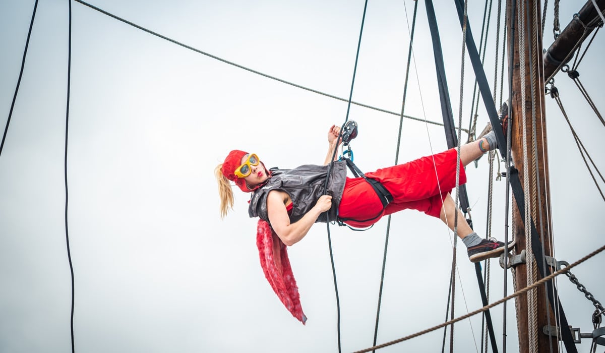 En kvinna som är cirkusartist hänger i ett rep och en sele med fötterna mot masten på ett segelfartyg.