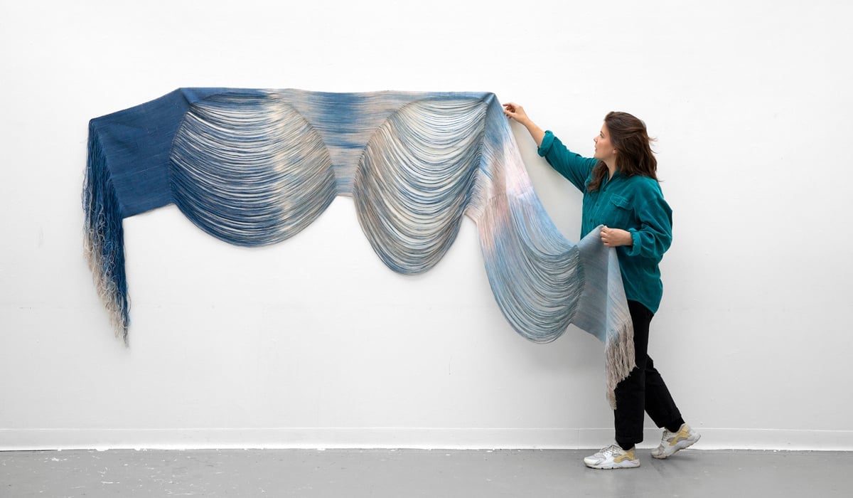 En kvinna håller i ett stort mjukt böljande blått tyg som är upphängt på en vit vägg