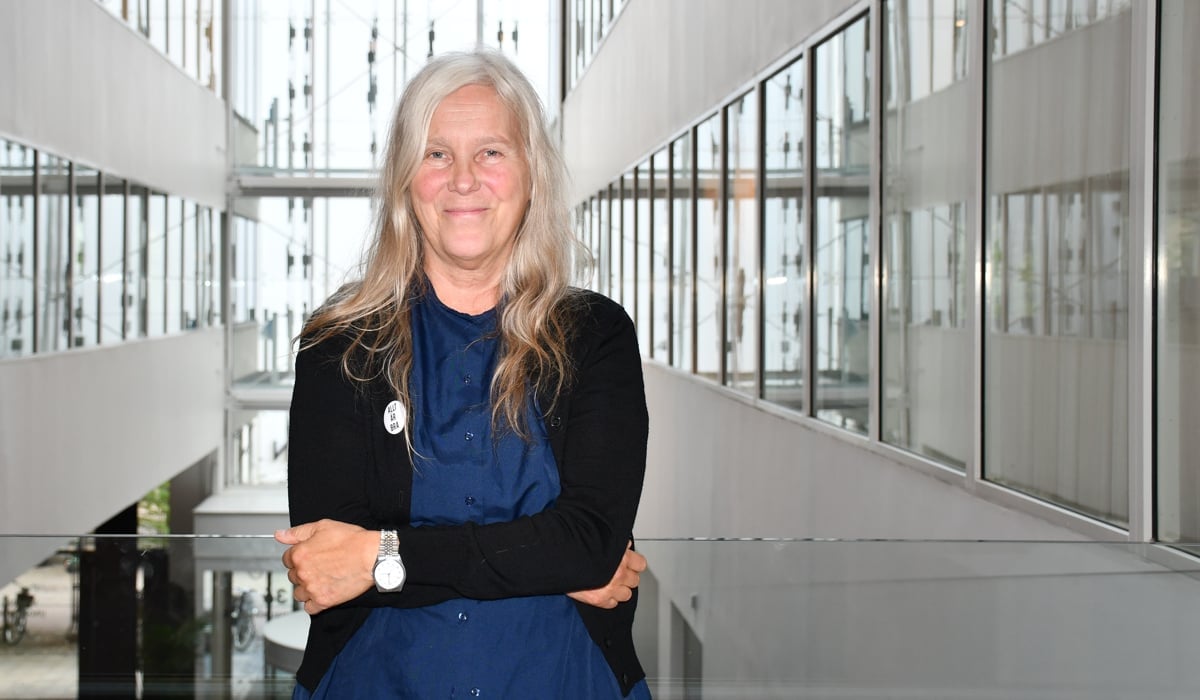 Marja-Leena ler mot fotografen med armarna löst runt midjan. Bakom stora glasfönster i kontorsbyggnad.