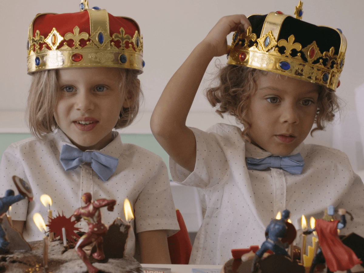 Två barn i kungakronor sitter vid ett bord där två födelsedagstårtor har tända ljus.