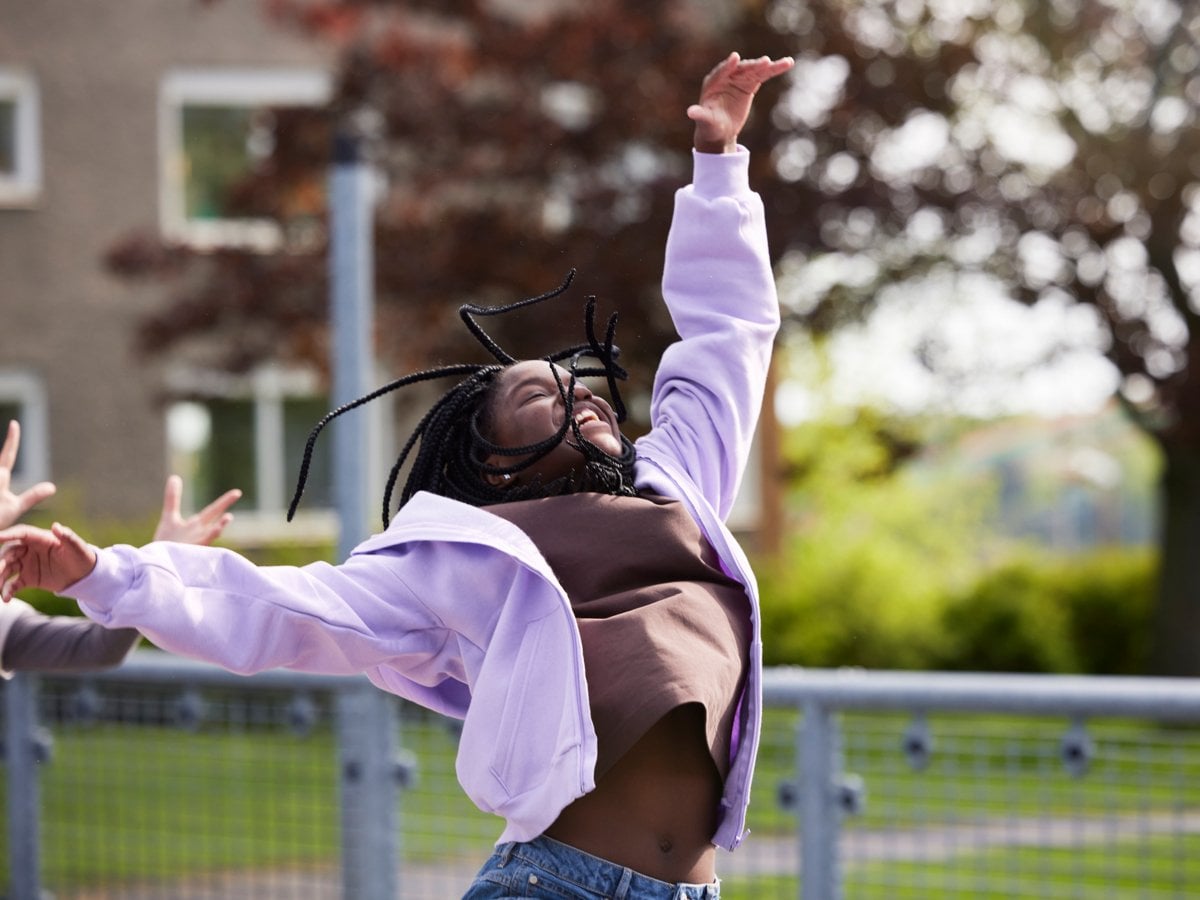 Flicka i brun tröja och lila munkjacka hoppar upp i luften och skrattar. I bakgrunden syns träd och lägenhetshus,