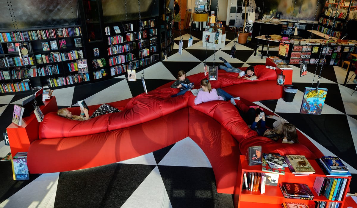 En stor röd soffa i ett rum fyllt med massor av böcker där barn ligger och sitter och läser.