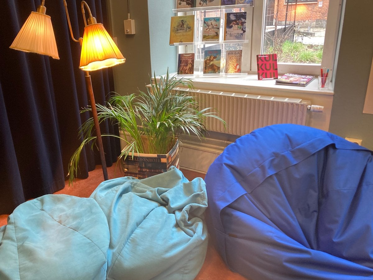 Bild på läshörna på Hornstulls bibliotek. Blåa sittpuffar, en krukväxt och en golvlampa framför ett fönster med böcker.