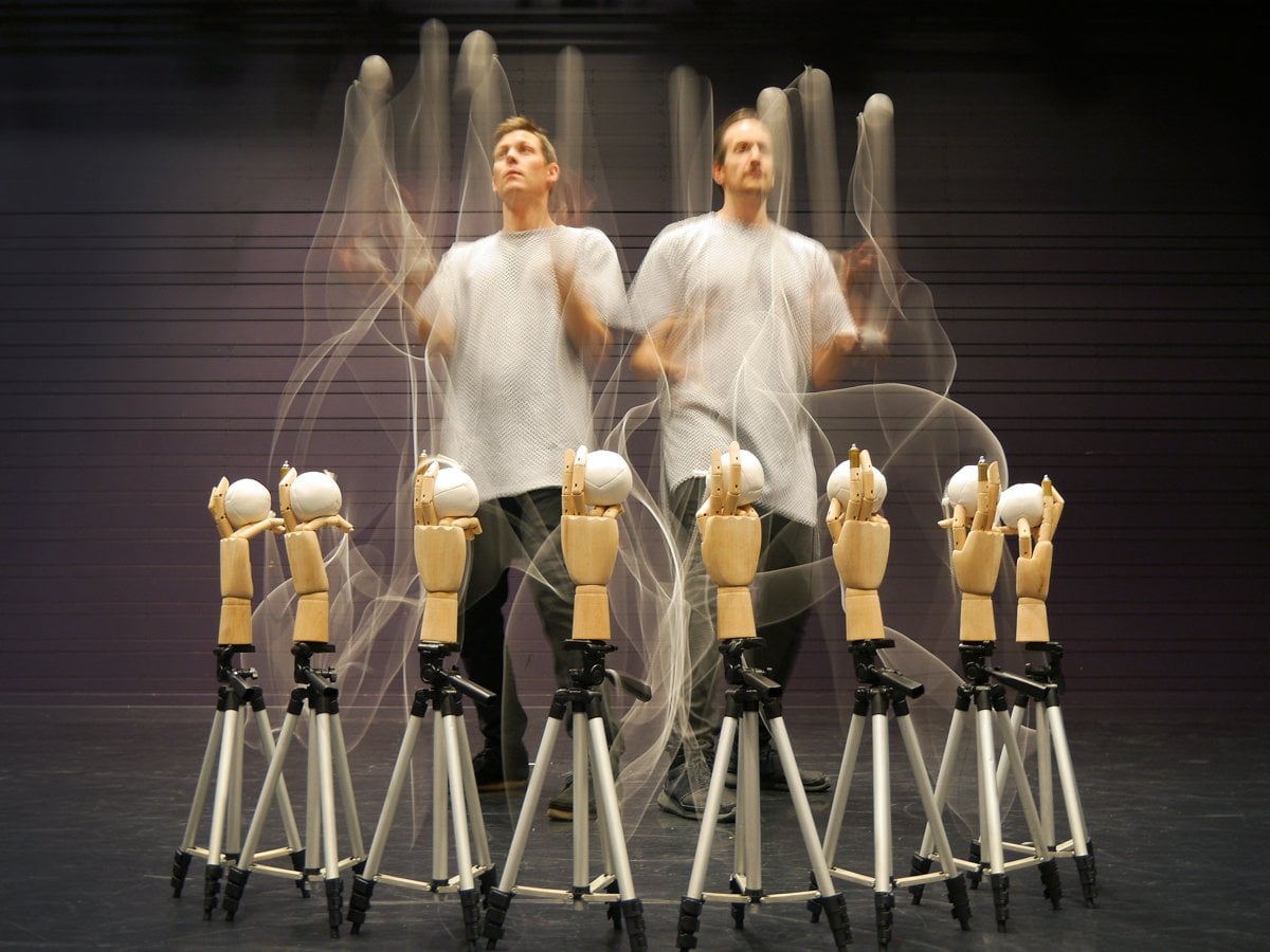 Två män jonglerar med vita bollar. Runtom dem en cirkel med stativ med händer av trä som håller i vita bollar.