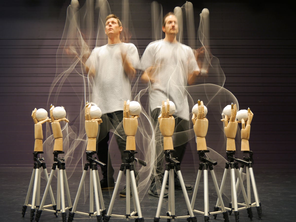 Två män jonglerar med vita bollar. Runtom dem en cirkel med stativ med händer av trä som håller i vita bollar.