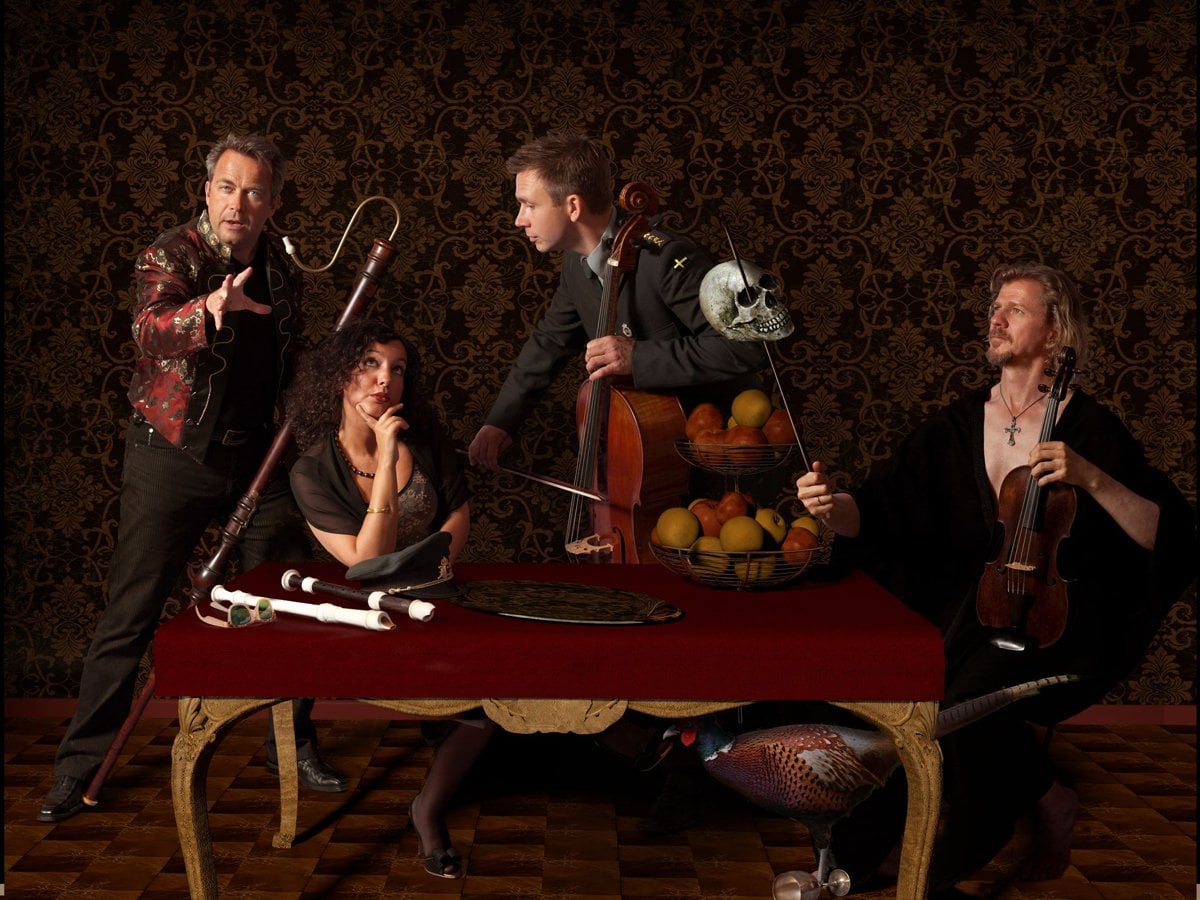 Fyra musiker framför en mönstrad tapet står med instrumenten bakom ett bord med frukter.