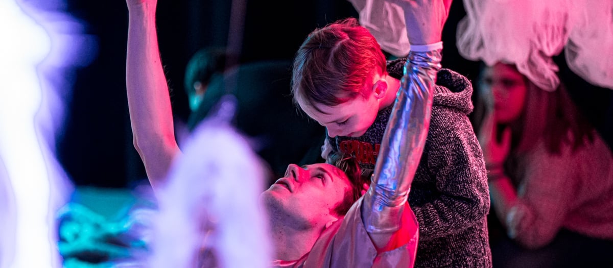 Ett barn står bakom en silverklädd dansare som sitter med armarna uppsträckta i luften. Runt om dem syns olika tylltyger.