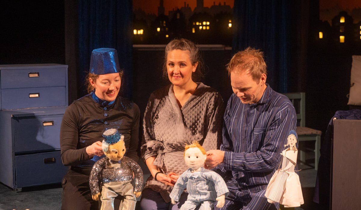 Tre dockteaterspelare på en scen med två dockor framför en scenografi föreställande en stad i skymning.