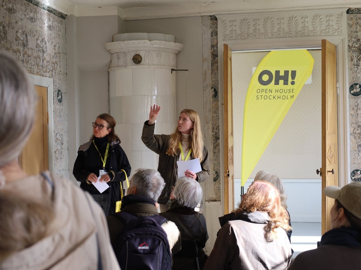 Två personer föreläser för en grupp åhörare i ett rum i äldre stil med kakelugn och mönstrade tapeter. Bakom talarna syns en vepa med texten Open House Stockholm.