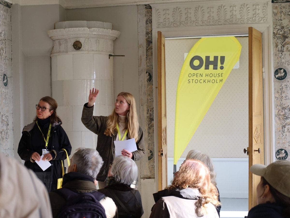 Två personer föreläser för en grupp åhörare i ett rum i äldre stil med kakelugn och mönstrade tapeter. Bakom talarna syns en vepa med texten Open House Stockholm.