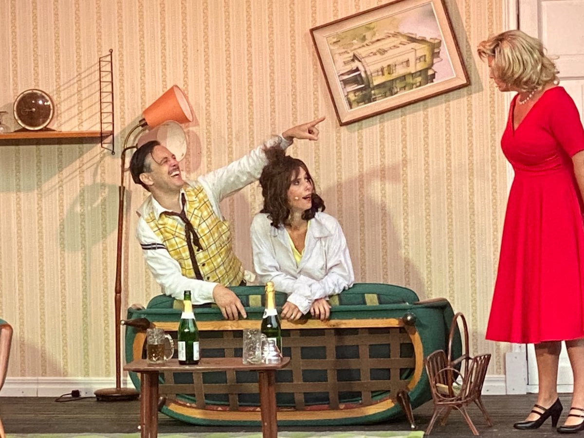 Tre skådespelare på scen. En man och en kvinna sitter i en soffa i en lägenhet. En kvinna i röd klänning står bredvid soffan och tittar på dem.