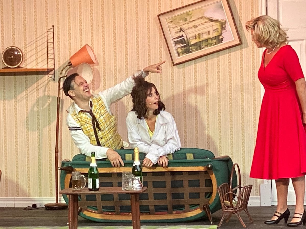 Tre skådespelare på scen. En man och en kvinna sitter i en soffa i en lägenhet. En kvinna i röd klänning står bredvid soffan och tittar på dem.